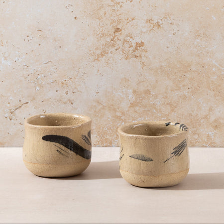 Two ceramic cups with feather designs on a beige surface and background