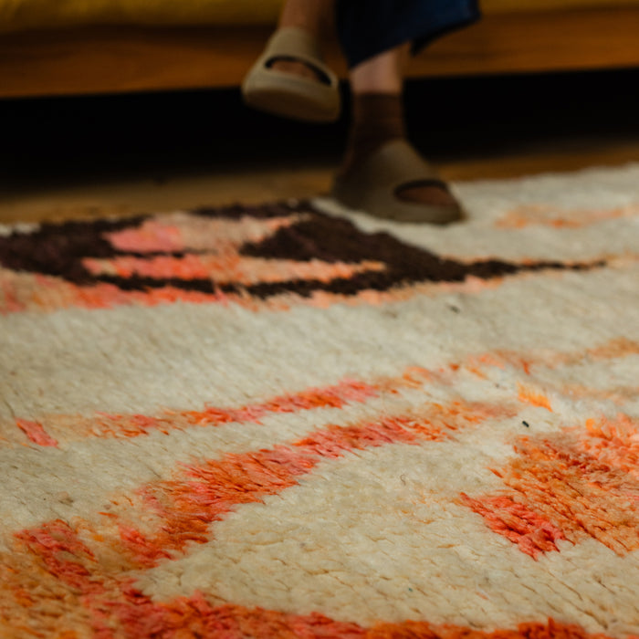 Moroccan Berber Boujad wool hand-knotted rug with abstract design in cream, pink and orange with brown. Someone is sitting nearby with sandals and feet on the rug.