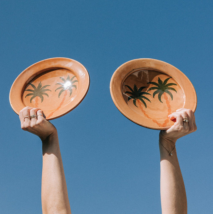 Two hands holding two terracotta plates with palm tree designs against a clear blue sky.