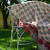 A chunky woven rug with a beige, brown, and red checkerboard pattern and fringe trim, draped over a white metal garden chair on a green grassy lawn under leafy trees.