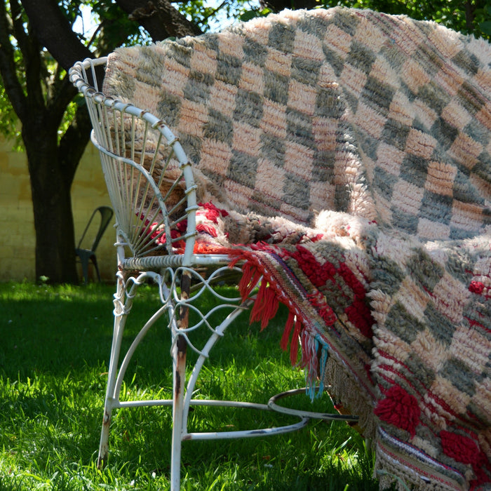 A chunky woven rug with a beige, brown, and red checkerboard pattern and fringe trim, draped over a white metal garden chair on a green grassy lawn under leafy trees.
