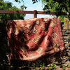 A red patterned wool rug with fringe is draped over a rustic stone wall in a sunny garden setting with trees and grass.