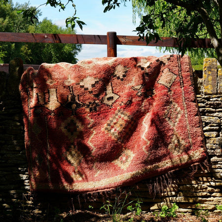 A red patterned wool rug with fringe is draped over a rustic stone wall in a sunny garden setting with trees and grass.