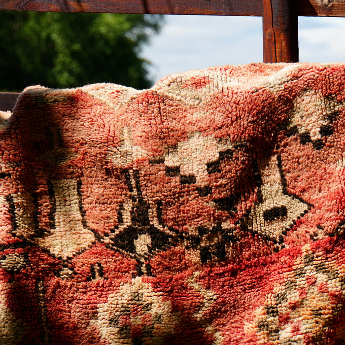 A close-up of a textured, patterned kilim rug draped over a dark wooden fence outdoors, with green trees and blue sky visible in the background.