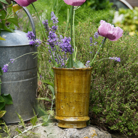 A rustic yellow glazed ceramic planter pot holding pink poppy flowers and purple lavender, styled outdoors on a stone ledge next to a galvanized metal watering can in a garden setting.