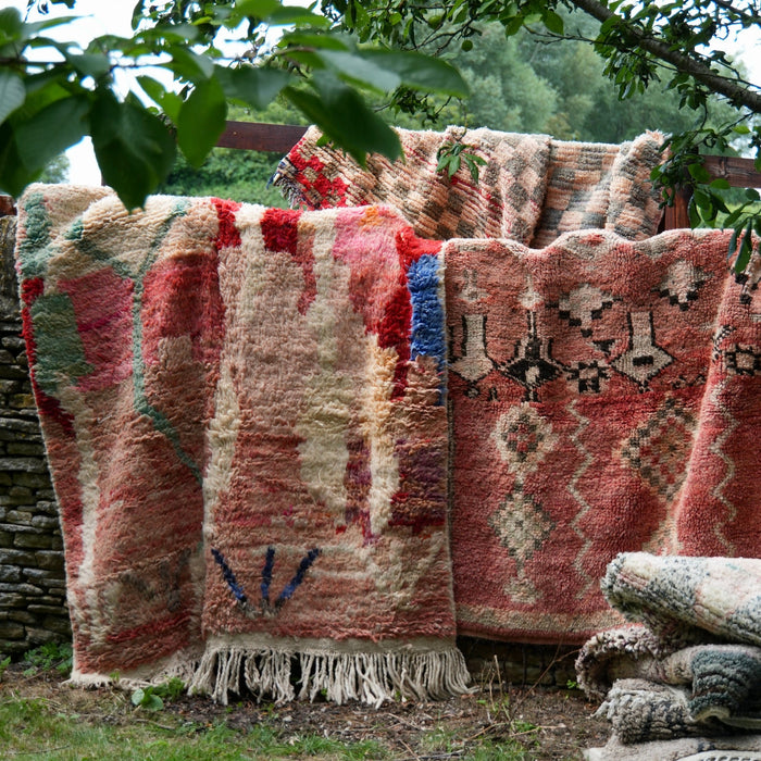 Several woven rugs and throws in earthy red, beige, brown, and muted green tones are draped outdoors on a wooden fence in a grassy garden with tree leaves overhead.