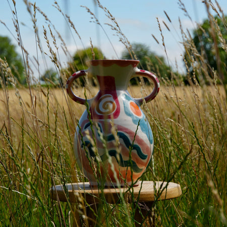 A hand-painted ceramic vase with two handles sits on a small wooden stool in a grassy field under a blue sky.