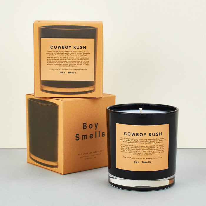 Boy smalls candle cowboy kush, black candle with yellow label on plain background with perfume boxes in orange. 