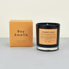 Boy smalls candle cowboy kush, black candle with yellow label on plain background with an orange perfume box next to the candle. 