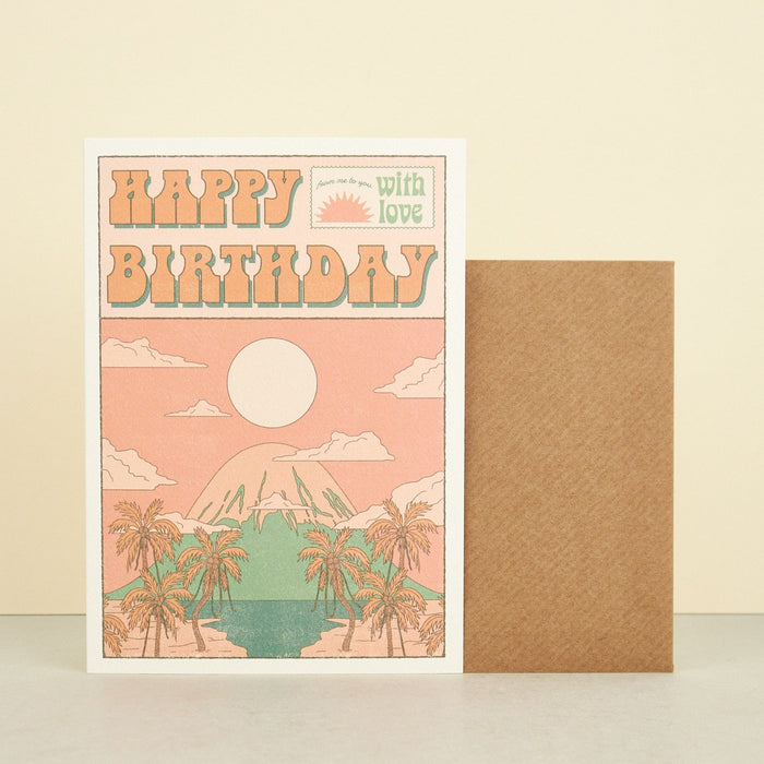 pink illustrated artisanal birthday card of a mountain with the sun and palm trees overlooking an ocean. 