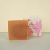 soap wrapped in beige paper with geometric pink and orange prints next to a bar of beige soapon a plain background. 