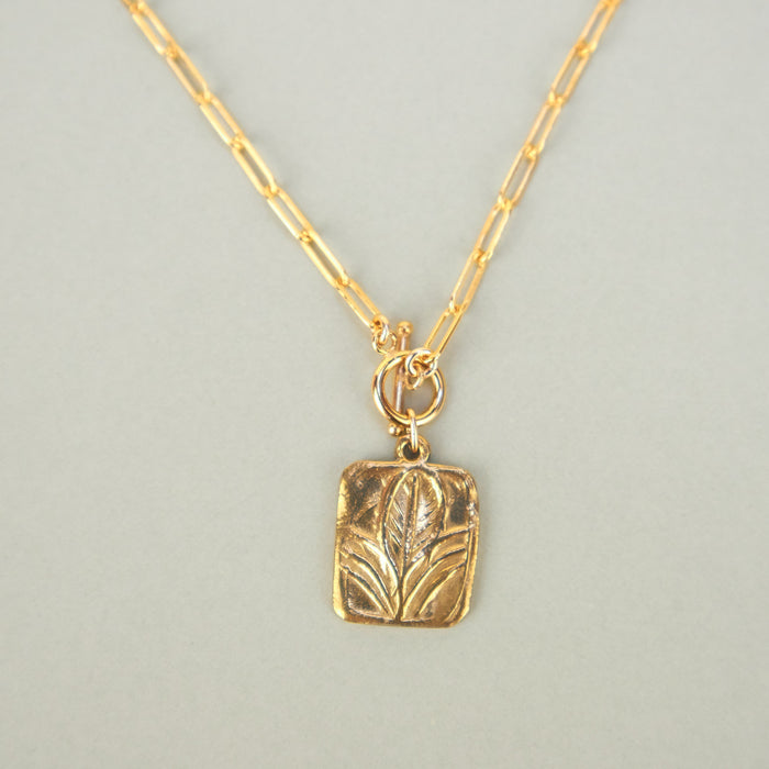 gold necklace with a square flat pendant of a leaf. plain background 