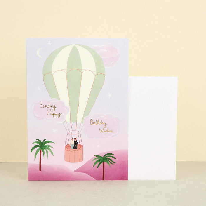 illustrated birthday card of a hot air balloon floating in a pink sky over a pink desert. a couple stand in the balloon. 